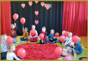 Dzieci siedzą wokół dużego serca. Trzymają czerwone balony.